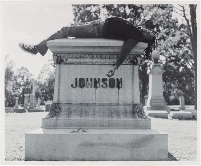 A Black man lying on top of a grave that reads 'JOHNSON.'