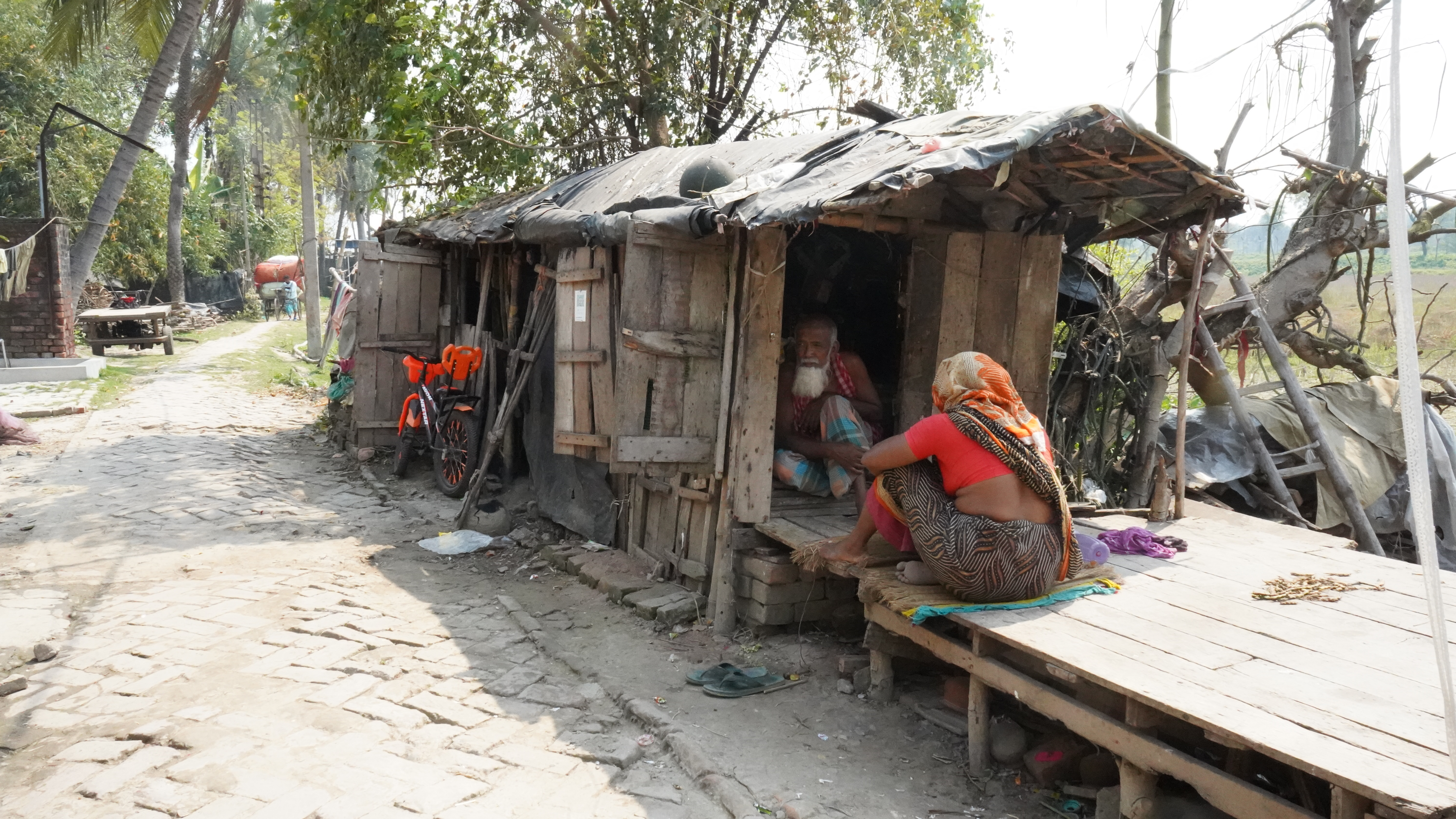 A woman sits on a wooden structure attached to a small building and a man sits in a doorway. The ground below is muddy with trees in the background.