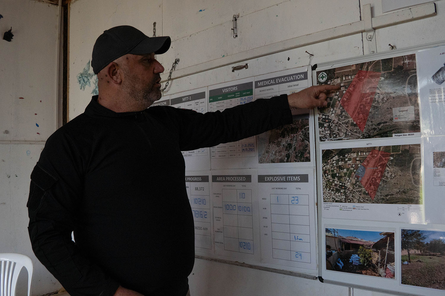 A man in a black shirt and black cap points at pictures and maps on the wall of a building.