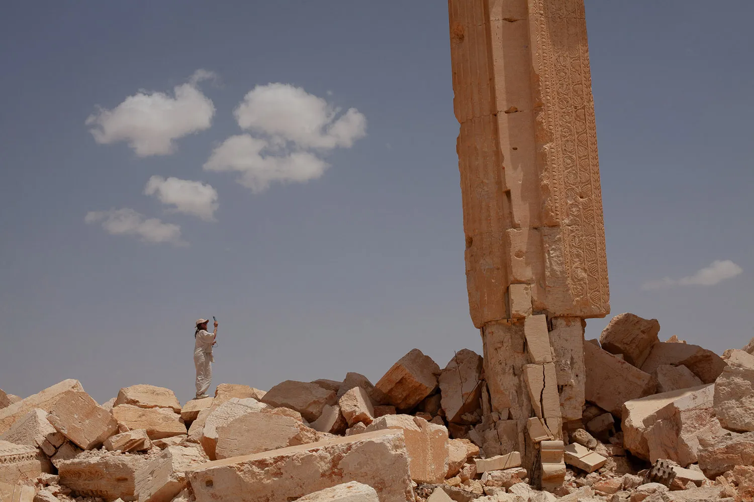 A large column looms over a small person standing atop a large field of rubble holding up a phone to take a photo.