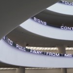 A partial view of a rotunda whose white wall is ringed with an LED screen displaying text. 'TO PULL AWAY FROM ME' is seen on one floor with doubled letters.