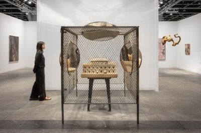 A woman walks behind a sculptural installation at an art fair booth. It consists of a cube of wired fencing with a three story palatial mansion on a metal pedestal at the center. A mirror above and one to the right and one to the left are cut into the cube and point at the center object.