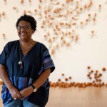A smiling Black woman in a blue shirt stands in front of an artwork installation.