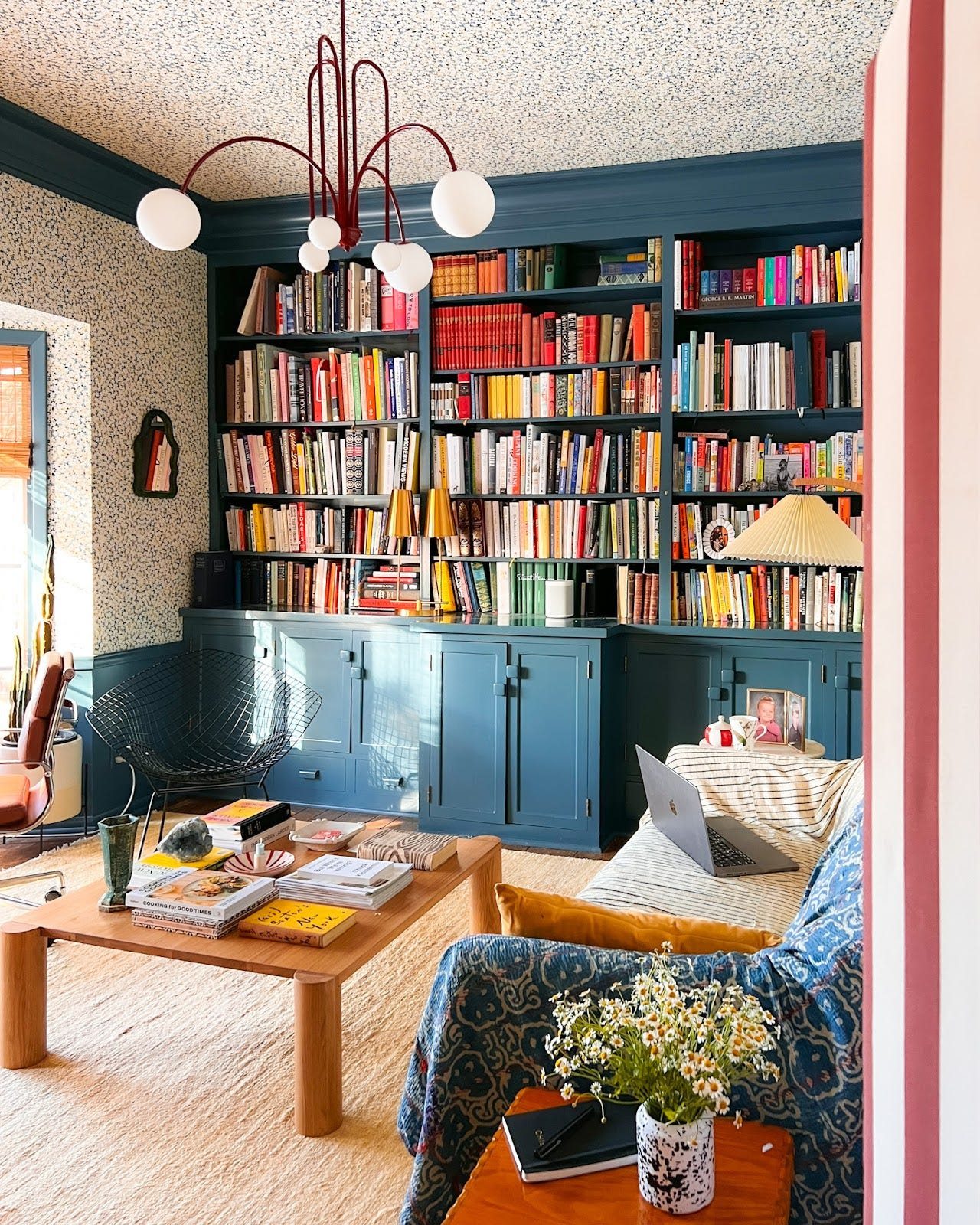 An office with a beige rug, small sofa, coffee table styled with books and decorative objects, floral wallpaper, and a blue built-in bookshelf filled with books