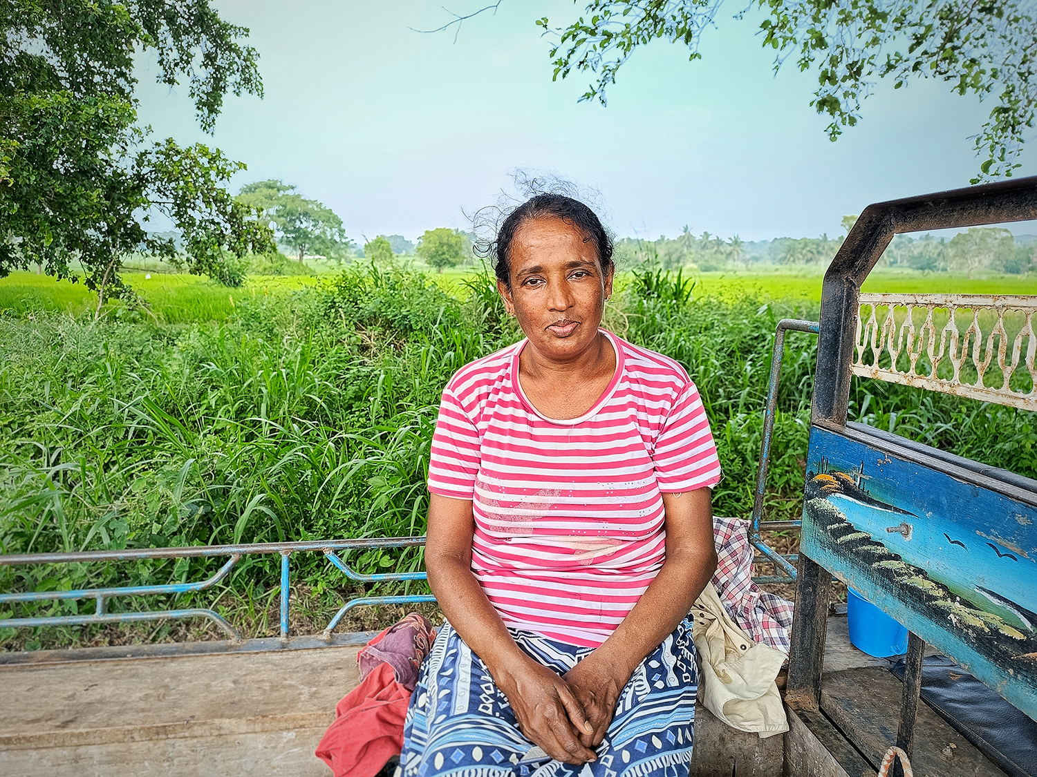 A woman in a striped shirt sits on the back of a farm vehicle with green fields behind her.