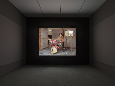 A screen in a darkened room showing a woman playing at a drum set.