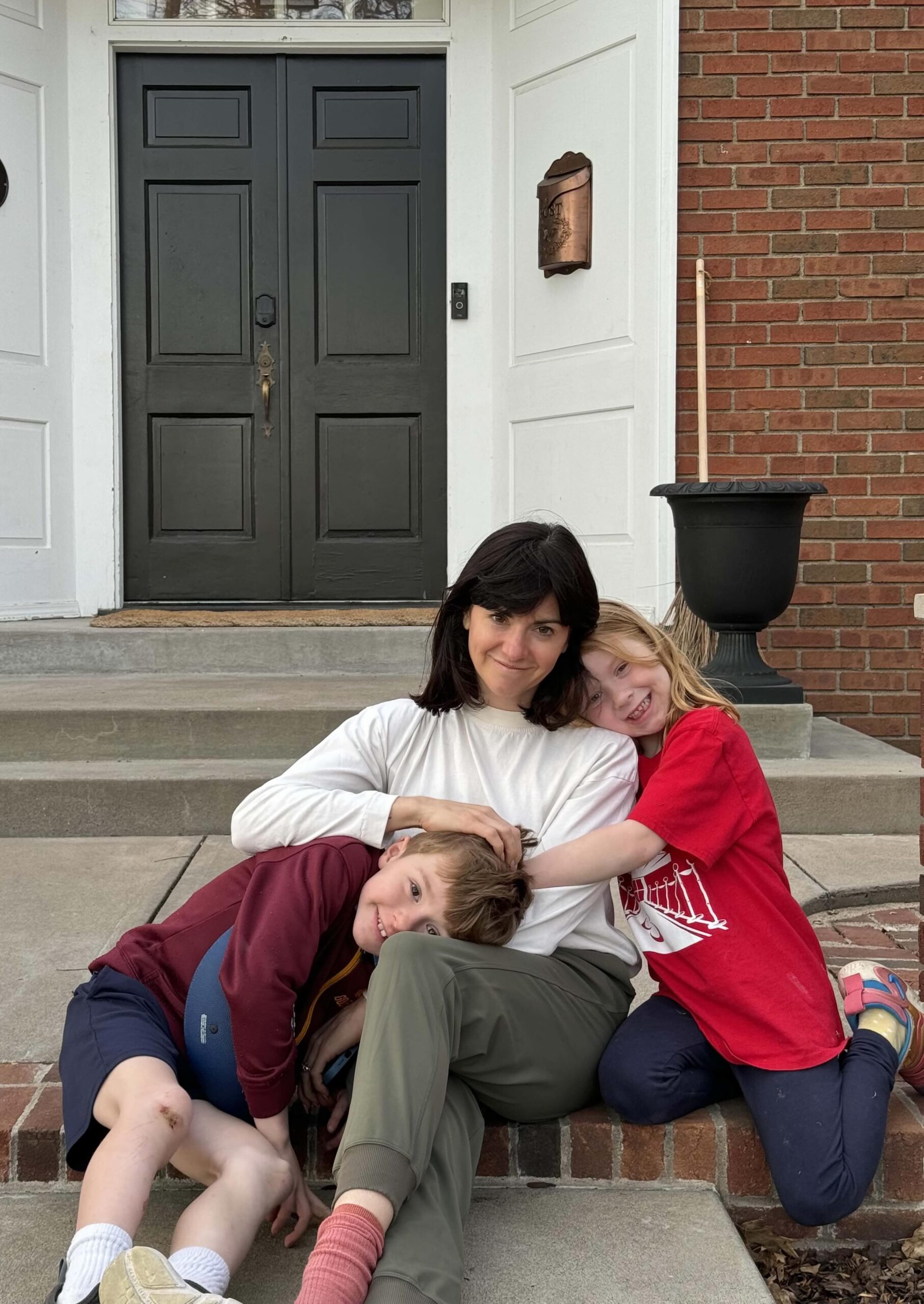 A mom and two kids sit on the front steps of their home, hugging and smiling at the camera