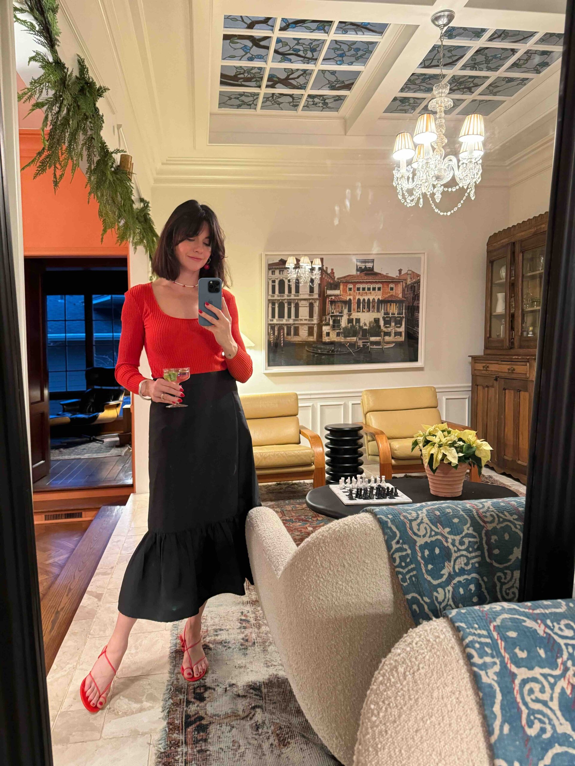 A woman wears a red sweater, long black skirt, and red sandals, holding a cocktail at a holiday party