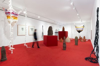 View of a gallery exhibition with sculptures on the floor and pedestals, as well as fiber works hanging from the ceiling and the walls. A blurred person walks in the center. 