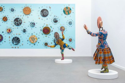 Two sculptures resembling faceless mannequins that raise their arms in front of a large painting.