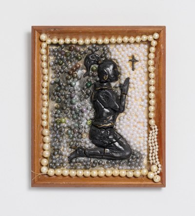 A mixed-media work, consisting of air dry clay, wood, Mod Podge, epoxy, acrylic, costume pearls, mother’s broken pearl necklace, showing a Black figure kneeling and praying to a small cross.