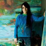 A white woman in a paint-splattered blue sweater placing one hand on an abstract canvas, with another canvas behind her.