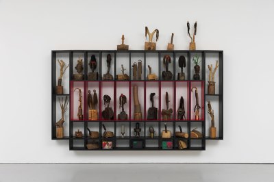A wall-hung sculpture that is divided into a cabinet-like structure with various plants and African masks in each section.
