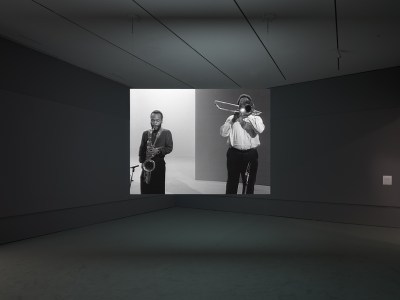 A hanging video screen showing two Black men, one playing the saxophone, the other playing a trombone.