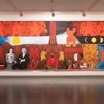 A large-scale painting showing a crouching red figure with breasts surrounded by a spread of other figures, moons, red scrawls, and more.