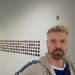 A young white man, artist Nick Dwyer, poses in an art gallery with a number of small images pinned to the wall behind him