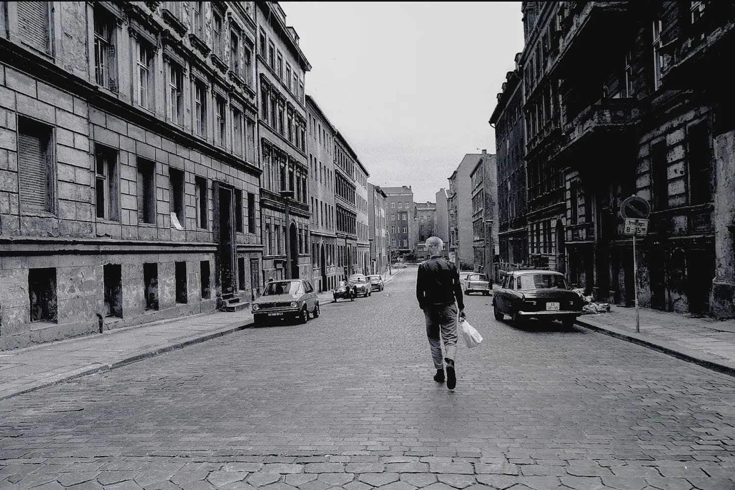 A person in a leather jacket walks down an empty street with buildings on either side.