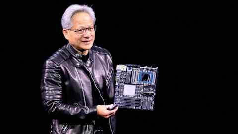 Jensen Huang holds up a circuit board while delivering a speech in a black leather jacket and standing against a dark background