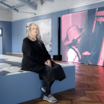 A woman seated on a platform in front of video monitors.