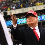 A man in a suit and a red hat tosses a coin at a stadium.