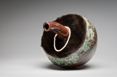 A ceramic sculpture with a spout. One end is covered with fur and a pearl bracelet hangs from the spout.