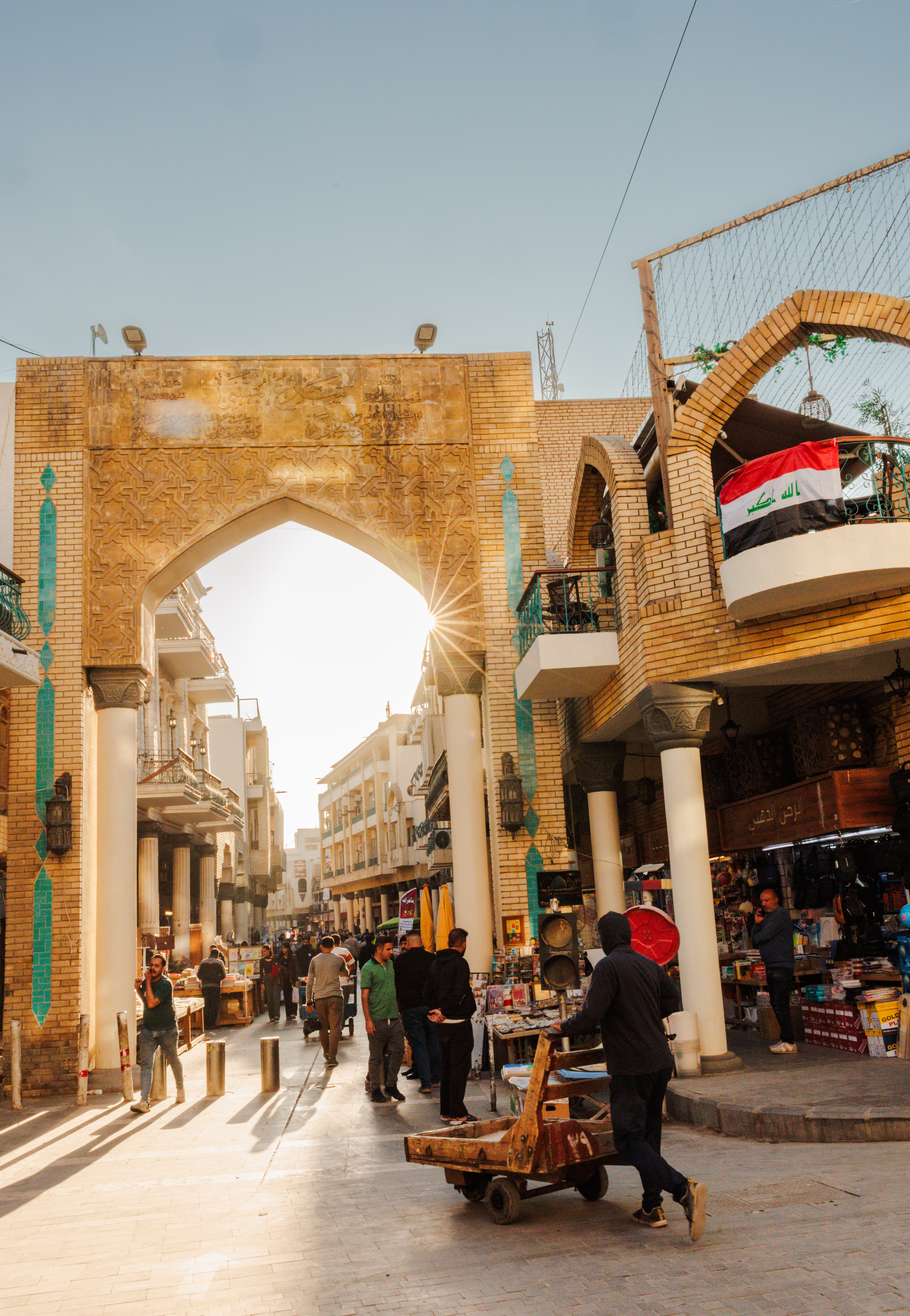 The sun glints through an archway the opens onto a busy street with people walking. In the foreground a person pushes a cart.