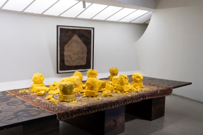 A table-like structure singed in places that has a Persian carpet laid on top, along with unevenly sized block of shea butter. Behind the table, there is a painting of a house.