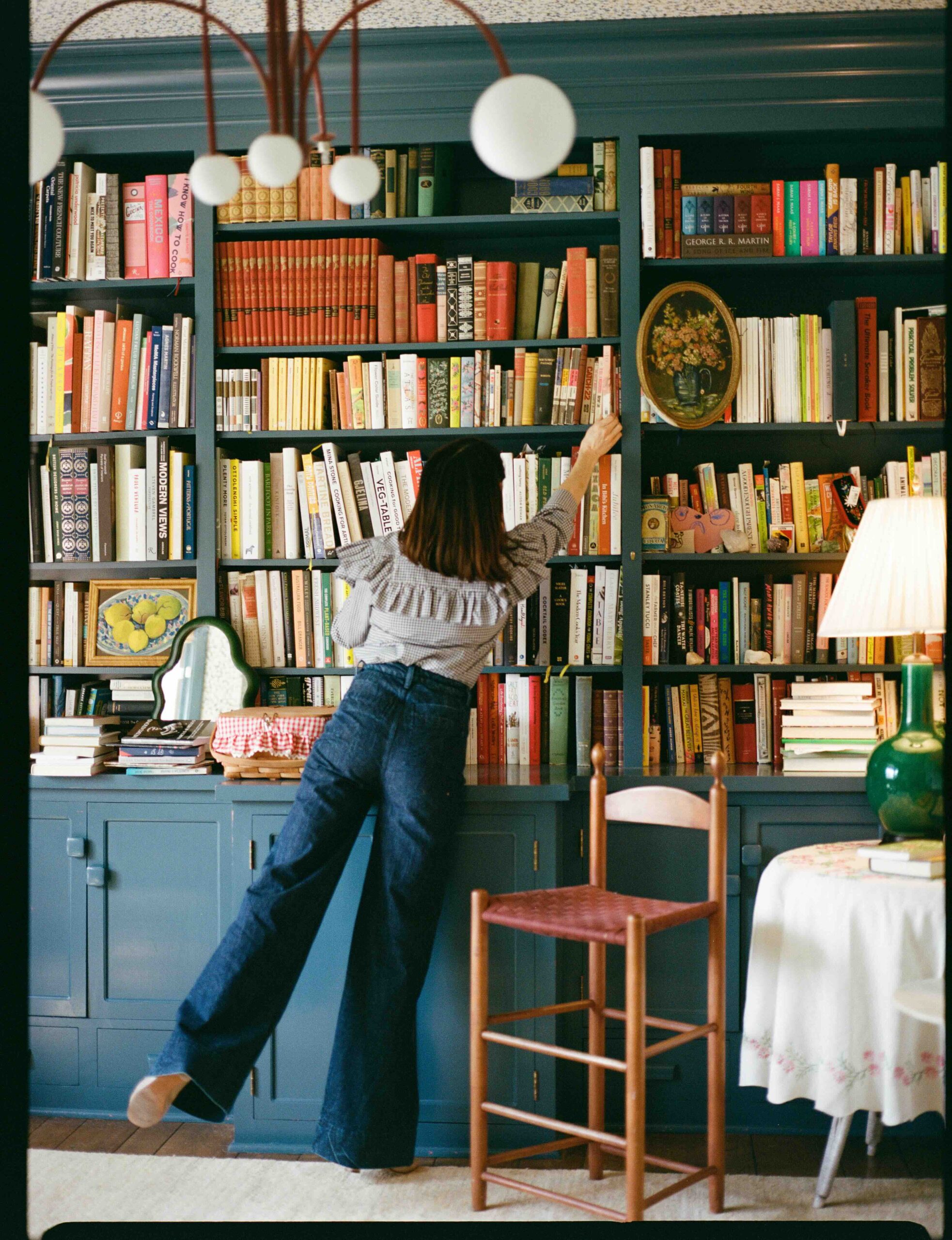 In a cozy home office, a woman stands in front of a blue bookshelf filled to the brim with books, reaching up to grab one from a top shelf