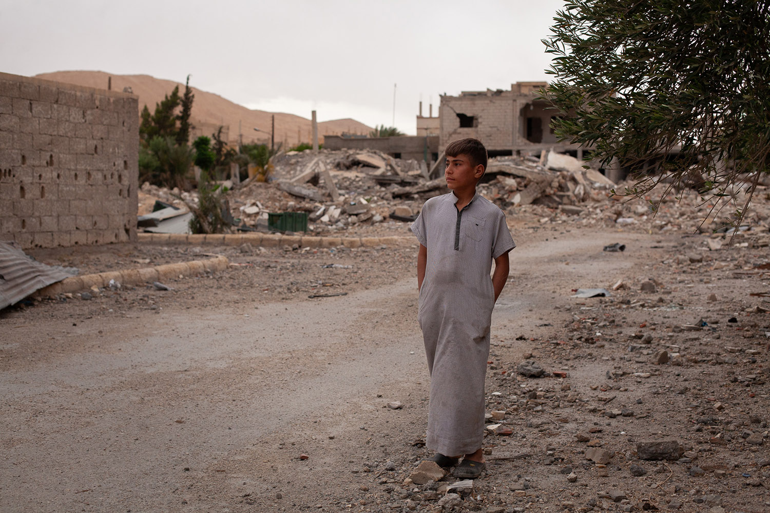 A boy with his hands in his pockets in a long robe on a dirt road covered with debris and damaged buildings behind.