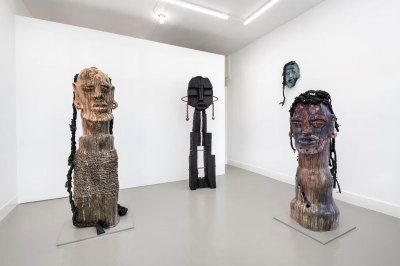 View of three large sculptures made of mixed media showing faces. One hangs at the wall behind.