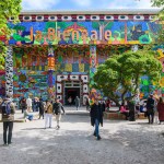 People walking in and out of a treed courtyard in front of a brightly painted building with columns. On its roof, there is the phrase 'la Biennale.'