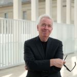 An older man in a black suit jacket and black shirt looks up with a slight smile as he holds black sunglasses in his right hand, with arms crossed. White architecture—many thin columns and a kind of fence stand behind him.