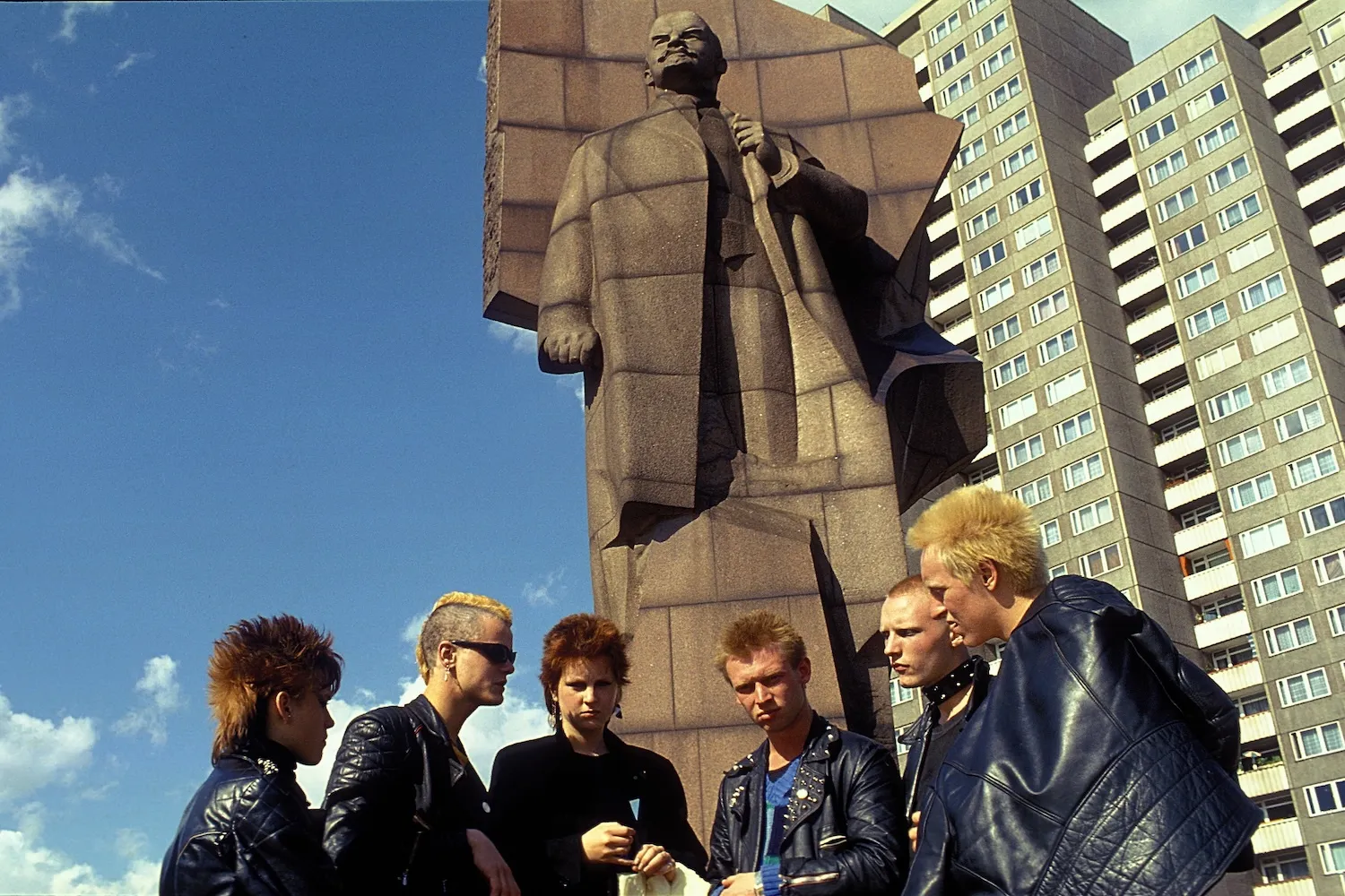Six youth in punk attire and haircuts stand in front of a statue of Lenin. Behind them is a tall apartment building.