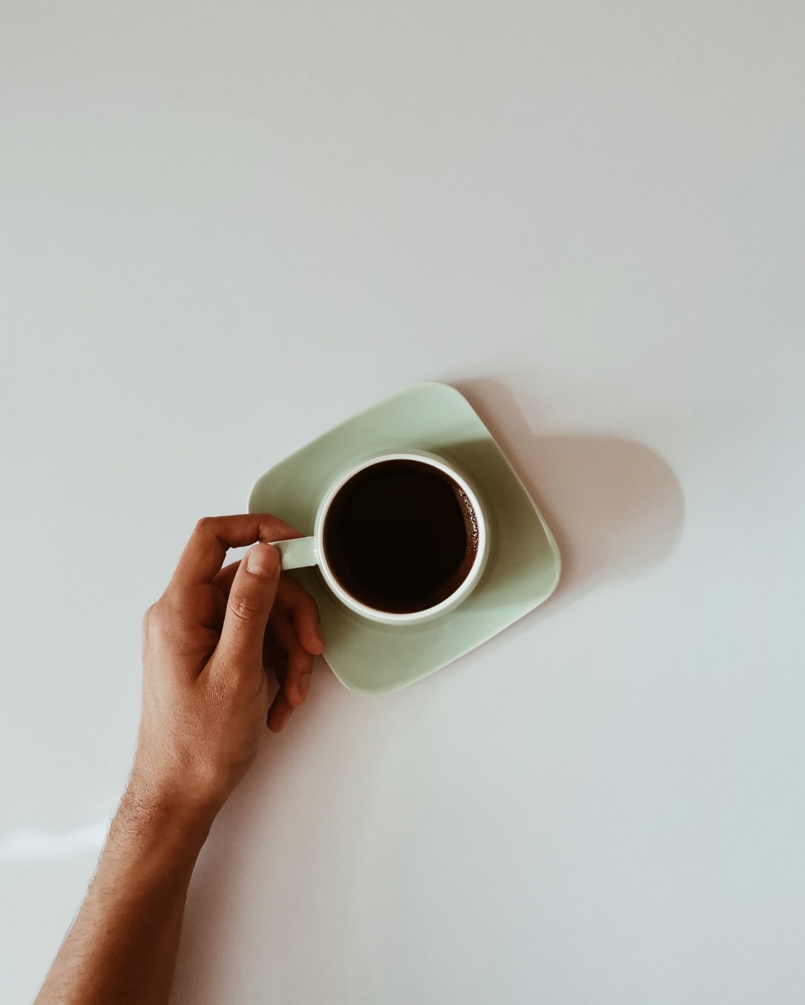 A seafoam green mug is filled with black coffee and sitting on a matching saucer. A person's hand is gently holding the handle of the mug. The scene is set against a white tabletop.