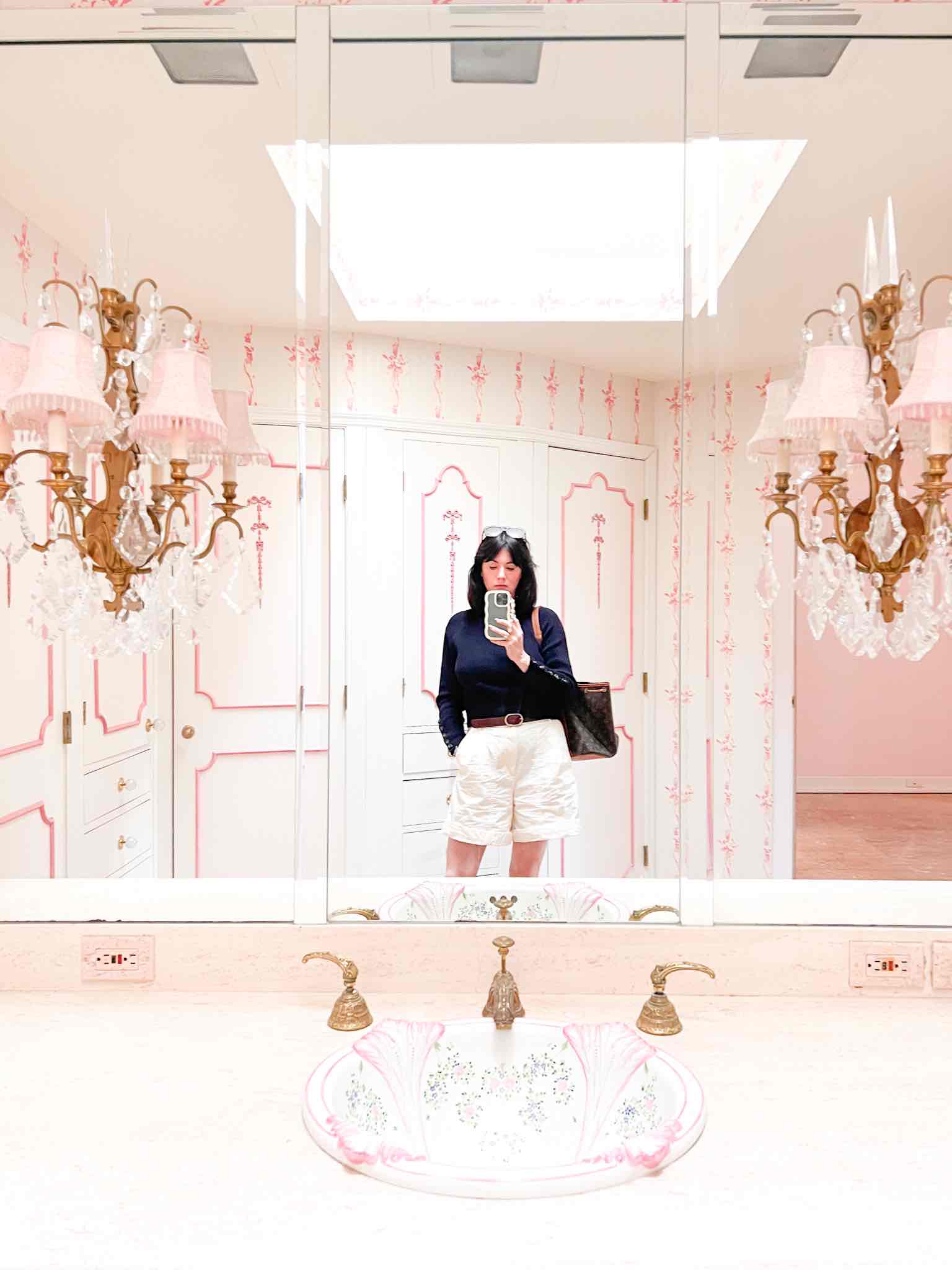 A woman wearing a navy sweater and white shorts stands in front of a mirror in a dated bathroom with pink and white decorative details galore