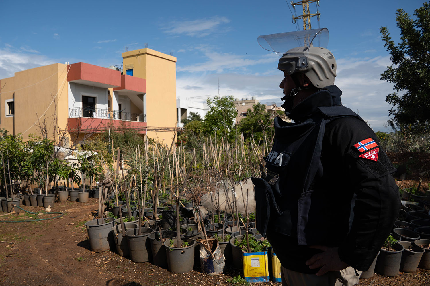A man in a protective coat, helmet, and mask looks toward a two-story stucco house. Plants in pots line a dirt road to the house.