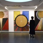 A woman staring at three large paintings with pyramidal forms topped with orbs.