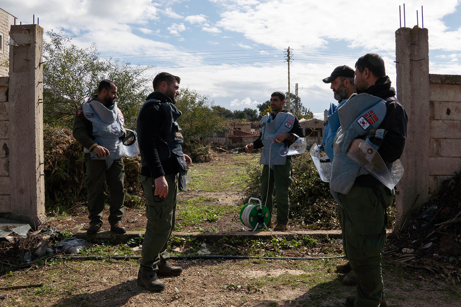 Five men in blue protective vests, with some holding helmets with visors, stand in the opening of a wall outdoors with low trees and blue sky behind them.