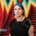 A woman with dyed-blue hair standing in front of a weaving featuring zigzagging lines in various colors.