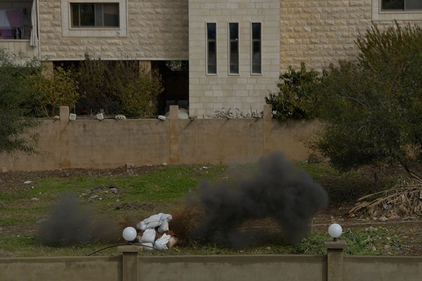 Smoke rises from a pile on the ground. A building is seen in the background.