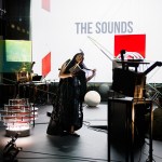 A room is filled with sculptures made of film projectors, as well has colorful squres that dangle in the space. A Black woman with long hair holds a book and wears a long dress as she appears to belt a song.