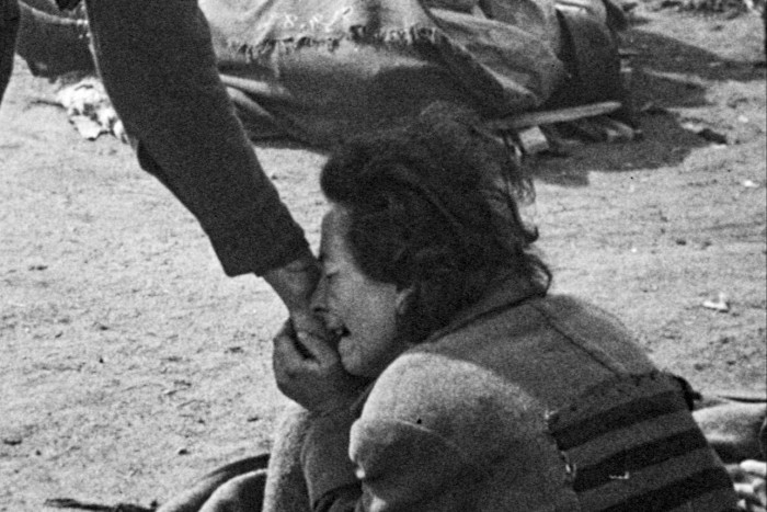 Crying woman kneeling on the ground clutches and kisses a person’s hand, wearing a striped uniform patch, surrounded by dirt and scattered belongings