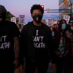 A Black man stands in the center of a color photograph. He wears a face mask and a T-shirt that says "I can't breathe."