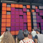 People gaze at a 25-foot tall installation made of woven squares in red, blue, and gold.