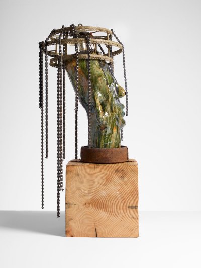 A sculpture of green-glazed ceramic head with a chandelier-like structure with chains on its head. It sits on a wooden base.