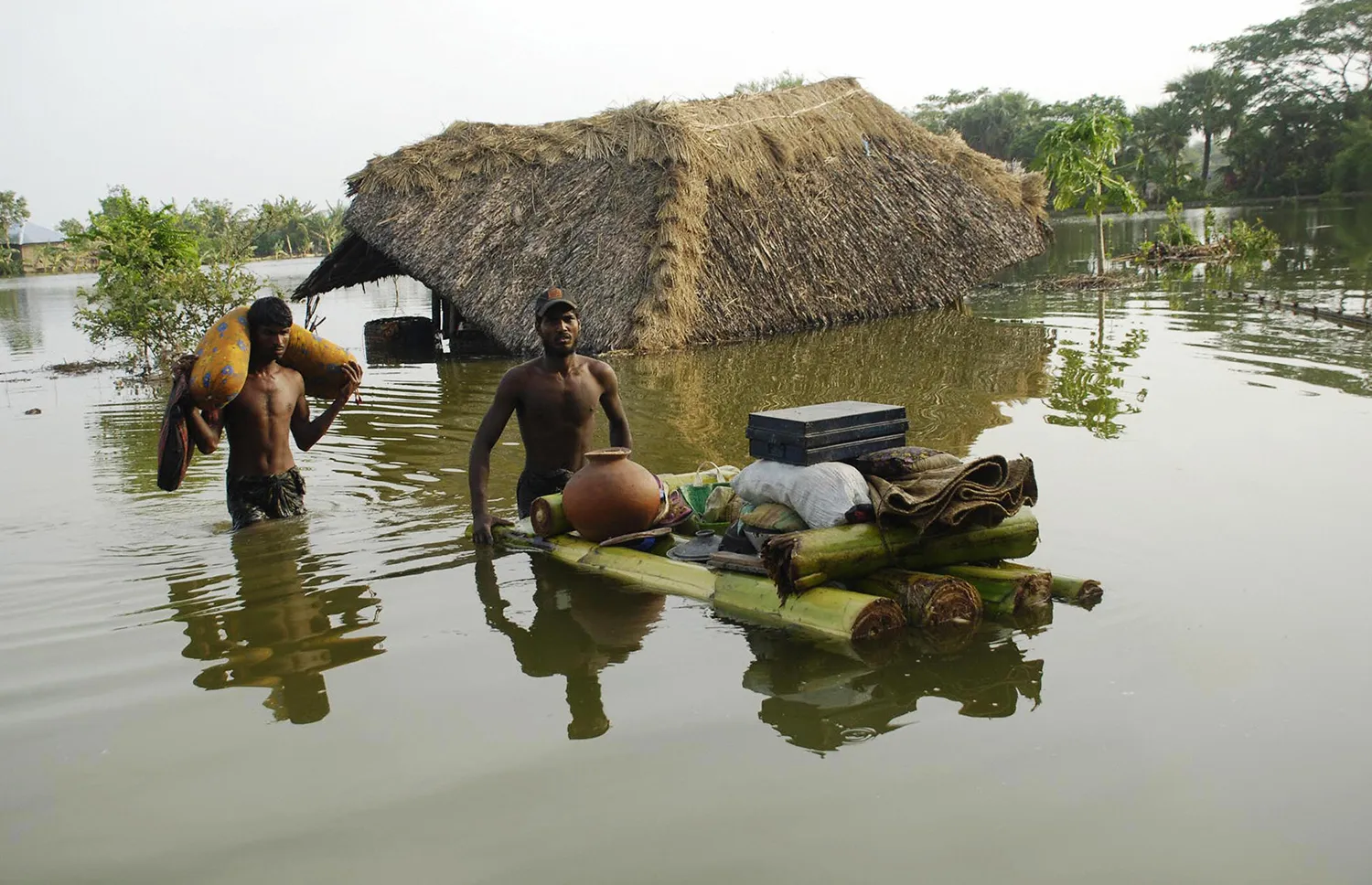 Two men carry their belongings, one pushing a laden raft through water, with a thatched building in the background.